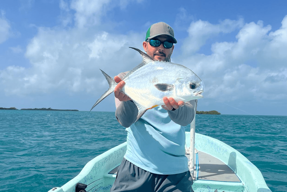 A man in a boat holds a shiny fish against a backdrop of clear blue water and sky.
