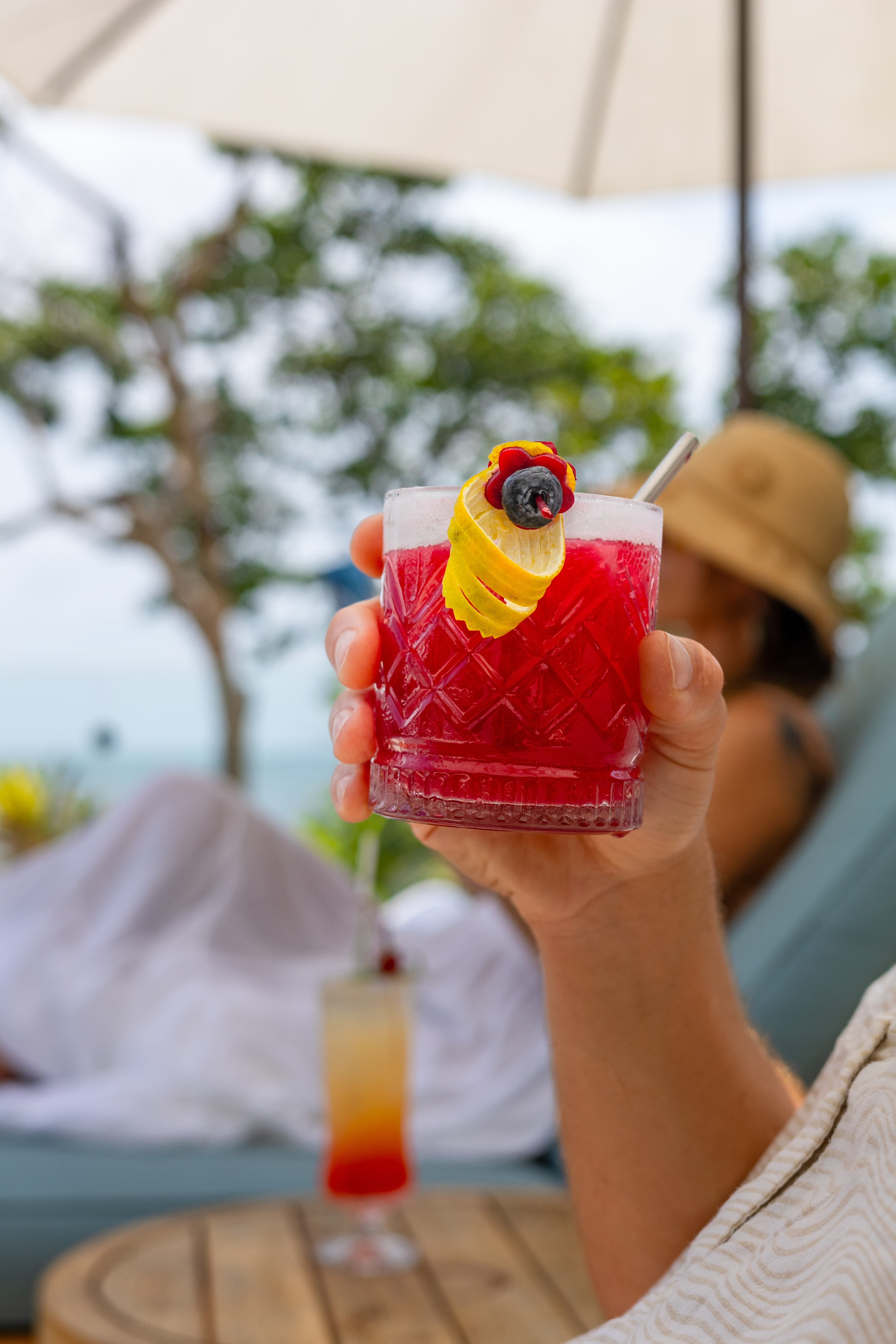 A hand holds a vibrant red cocktail garnished with a yellow twist and a berry, with a blurred beach scene in the background.