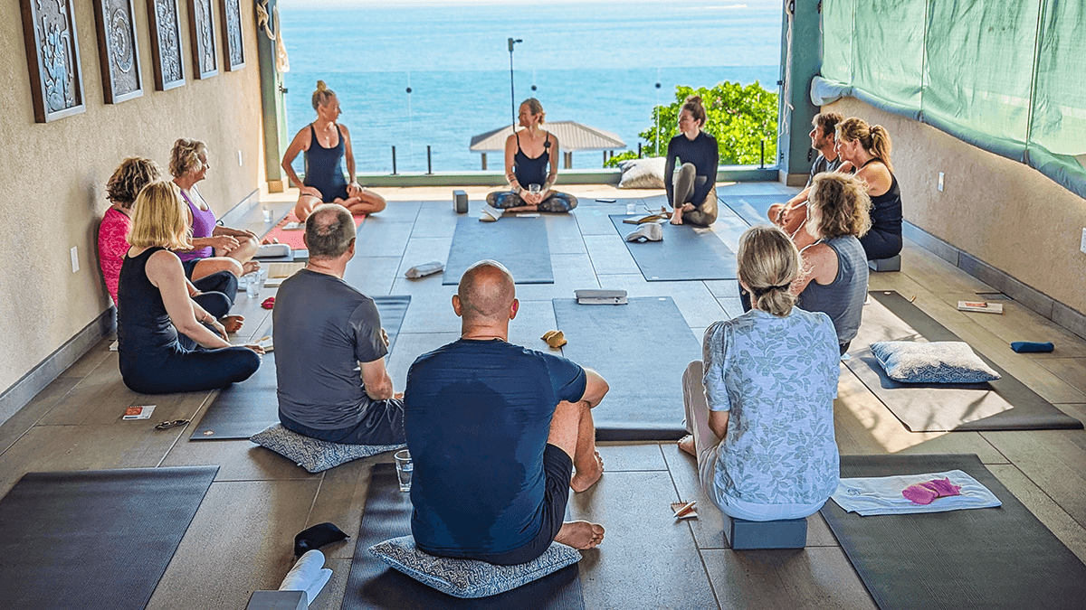 A group of people participates in a yoga class in a bright space overlooking the ocean.