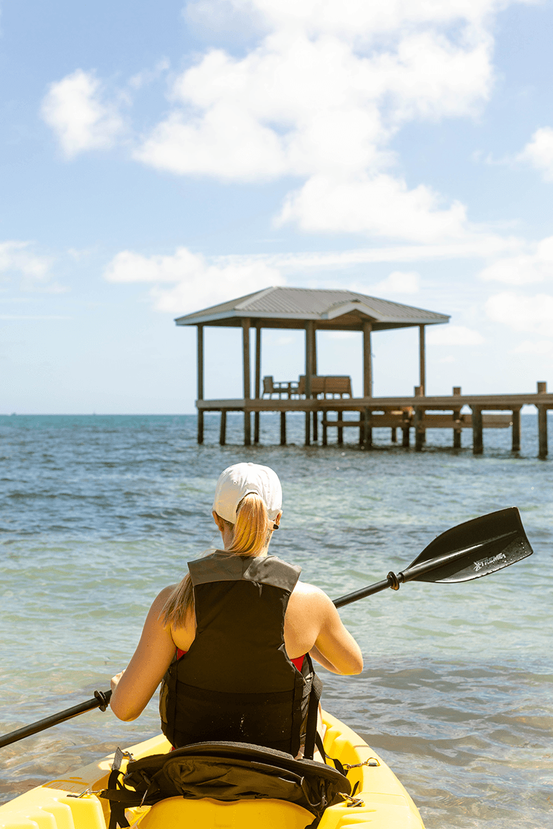 A person kayaking in calm waters, facing a wooden dock under a blue sky.