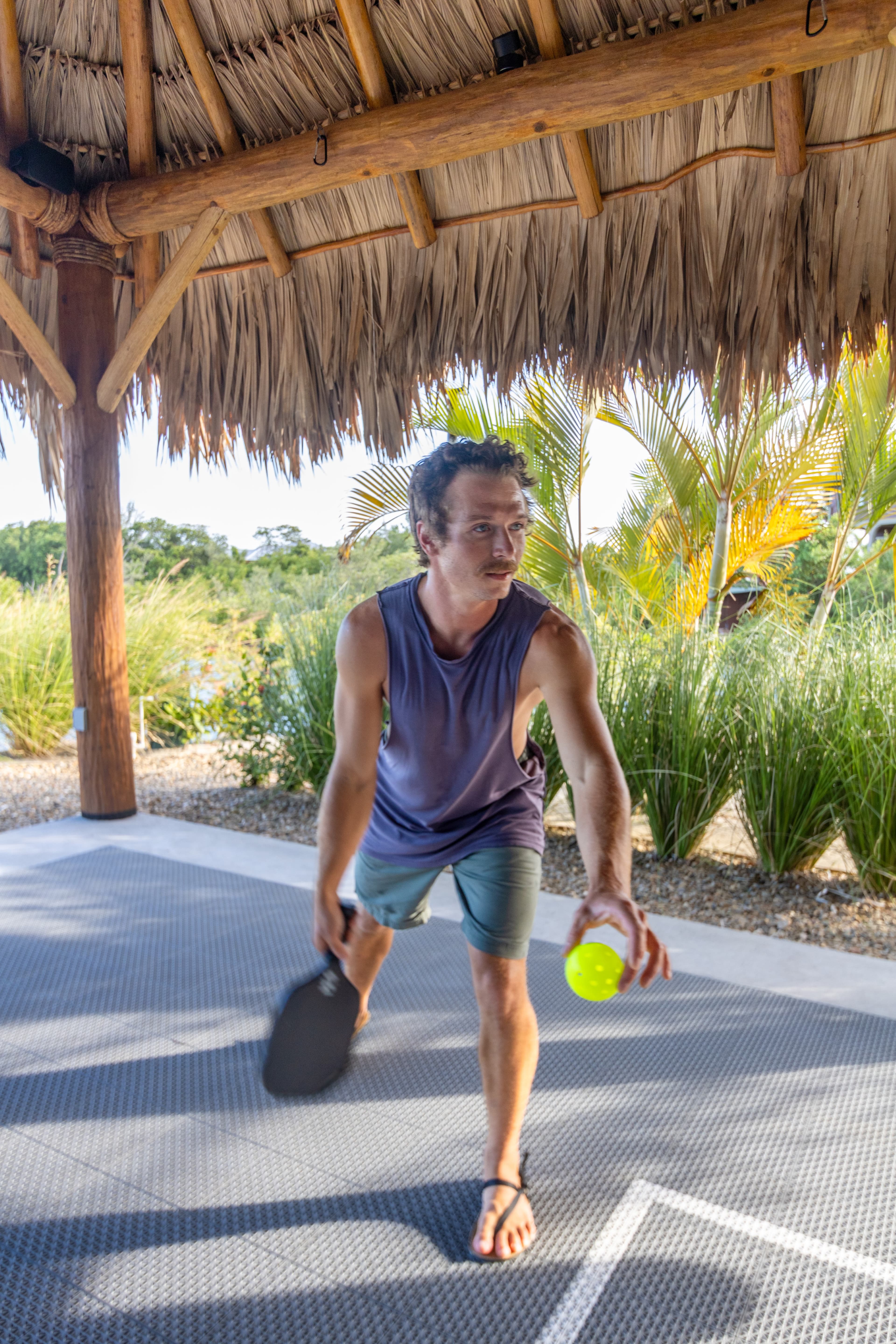 A man prepares to strike a ball while playing pickleball under a thatched roof.