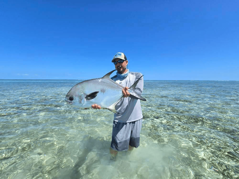 A man stands in shallow water holding a large fish, with a clear sky and ocean in the background.